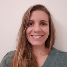 The image is a portrait of a woman with a warm smile. She has long, straight light brown hair, parted to one side. Her skin tone is fair, and her eyes are bright and welcoming. She is wearing a teal green top with a rounded neckline. The background appears to be a plain, light-coloured wall. Her overall expression and demeanor suggest a friendly and approachable individual.