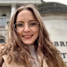 portrait of a woman smiling at the camera. She has long, wavy, light brown hair and is wearing thin-rimmed, round eyeglasses. The woman is dressed in a light turtleneck top, and what seems to be a brown coat over it. The background is blurred but suggests an indoor setting, possibly a café or a similar casual environment with warm lighting. She looks to be in a cheerful mood, captured in a moment that seems casual and genuine.