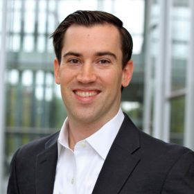 Man with a friendly expression. He is wearing a dark suit jacket over a white button-up shirt. The background suggests an indoor setting, possibly a professional environment like an office or a corporate building. The person is well-groomed with short dark hair, and he is looking directly at the camera, giving a warm and approachable appearance.