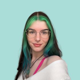 This image depicts a young woman with glasses, looking directly at the viewer. She has long hair with a gradient of colours, transitioning from a deep green at the roots to a vibrant turquoise at the tips. She wears a white off-the-shoulder top, complemented by a thin pink strap visible on one shoulder. The background is a soft, uniform teal, which subtly enhances the colours in her hair.