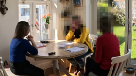 A bright, sunlit room where three individuals are seated around a wooden dining table. The setting appears informal, with notebooks and papers on the table suggesting a discussion or meeting. The room has large glass doors leading to a green garden, adding a welcoming and open atmosphere.